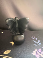 A small black butterfly-shaped figurine or ornament standing upright on a round black base, photographed against a dark fabric backdrop with a floral pattern visible on the surface.