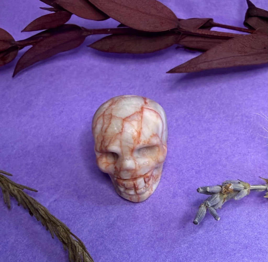 Crystal skull carving on purple background with feathers and botanicals — The Crooked Moon