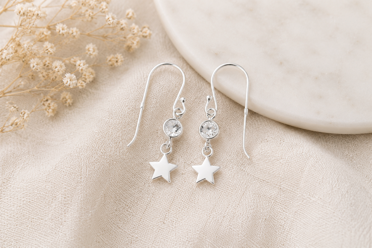A pair of silver dangle earrings with a small clear crystal and a star charm, displayed on a beige fabric surface next to dried flowers and a marble coaster.
