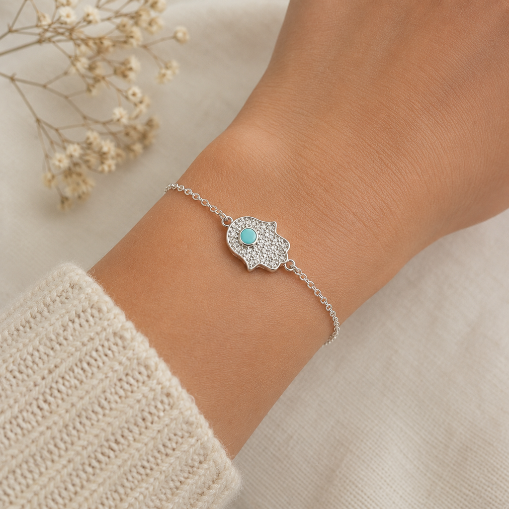 "Hamsa Hand Bracelet Sterling Silver worn shot — delicate silver chain bracelet with pavé CZ hamsa hand charm and turquoise stone shown worn on model's wrist over white knit sleeve, dried gypsophila in background"
