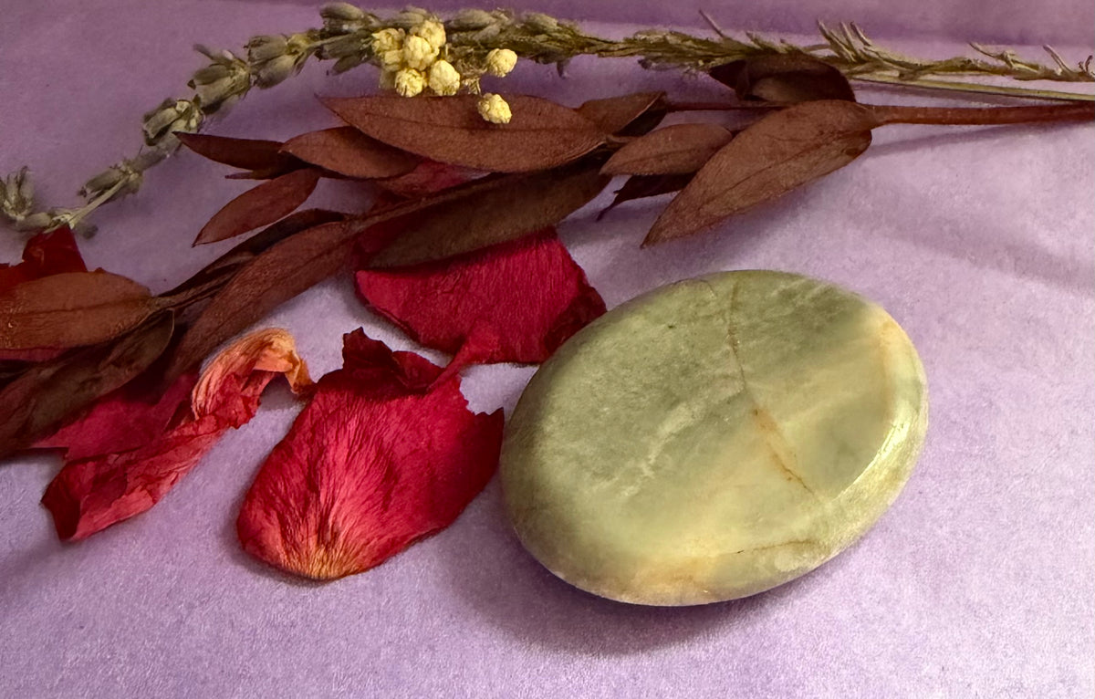 Moonstone palm stone placed on a lavender fabric surface, styled with dried brown leaves, red rose petals, and a dried flower sprig.