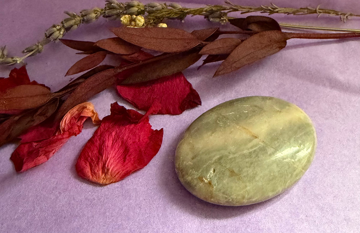 Moonstone palm stone with natural veining, styled on a lavender fabric background alongside dried brown leaves, red rose petals, and a sprig of dried lavender.