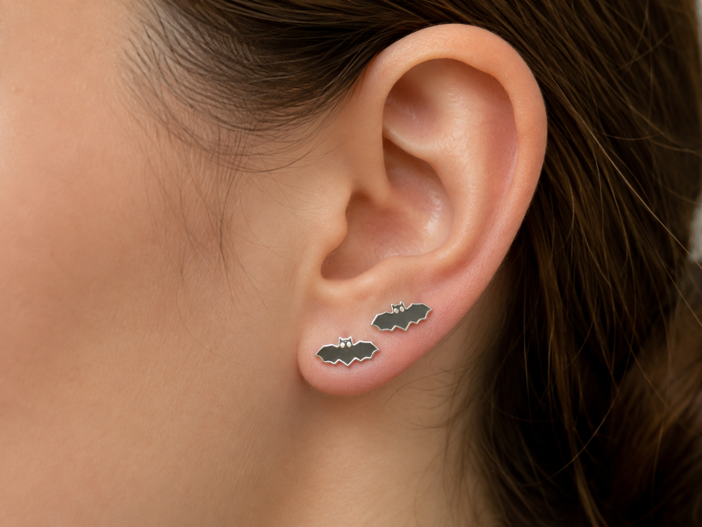 A close-up of a person's ear wearing two small bat-shaped stud earrings with dark gray enamel and silver outlines.