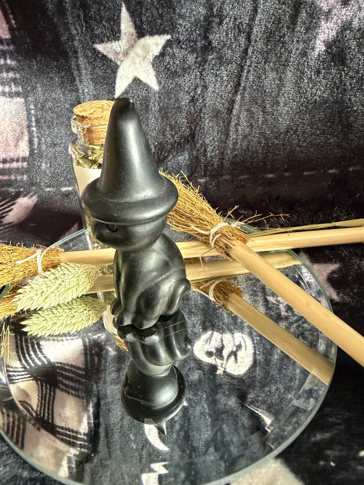 A small matte black wizard figurine with a pointed hat, displayed on a round glass tray with dried wheat and broomsticks against a dark patterned fabric backdrop.