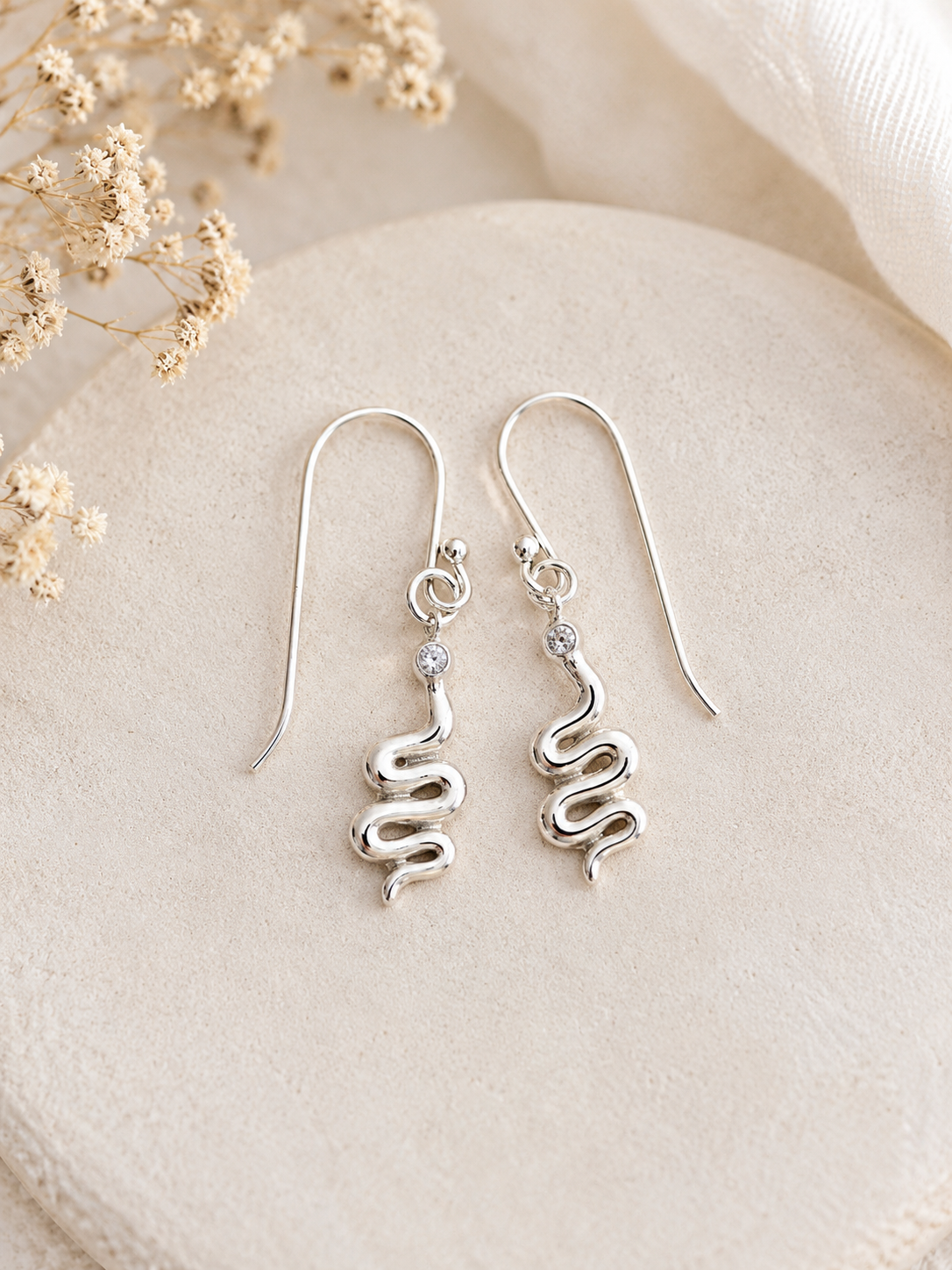 "Snake Drop Earrings Sterling Silver product shot — pair of polished silver hook earrings with coiled snake charm and CZ stone detail, on cream stone tray with dried gypsophila"