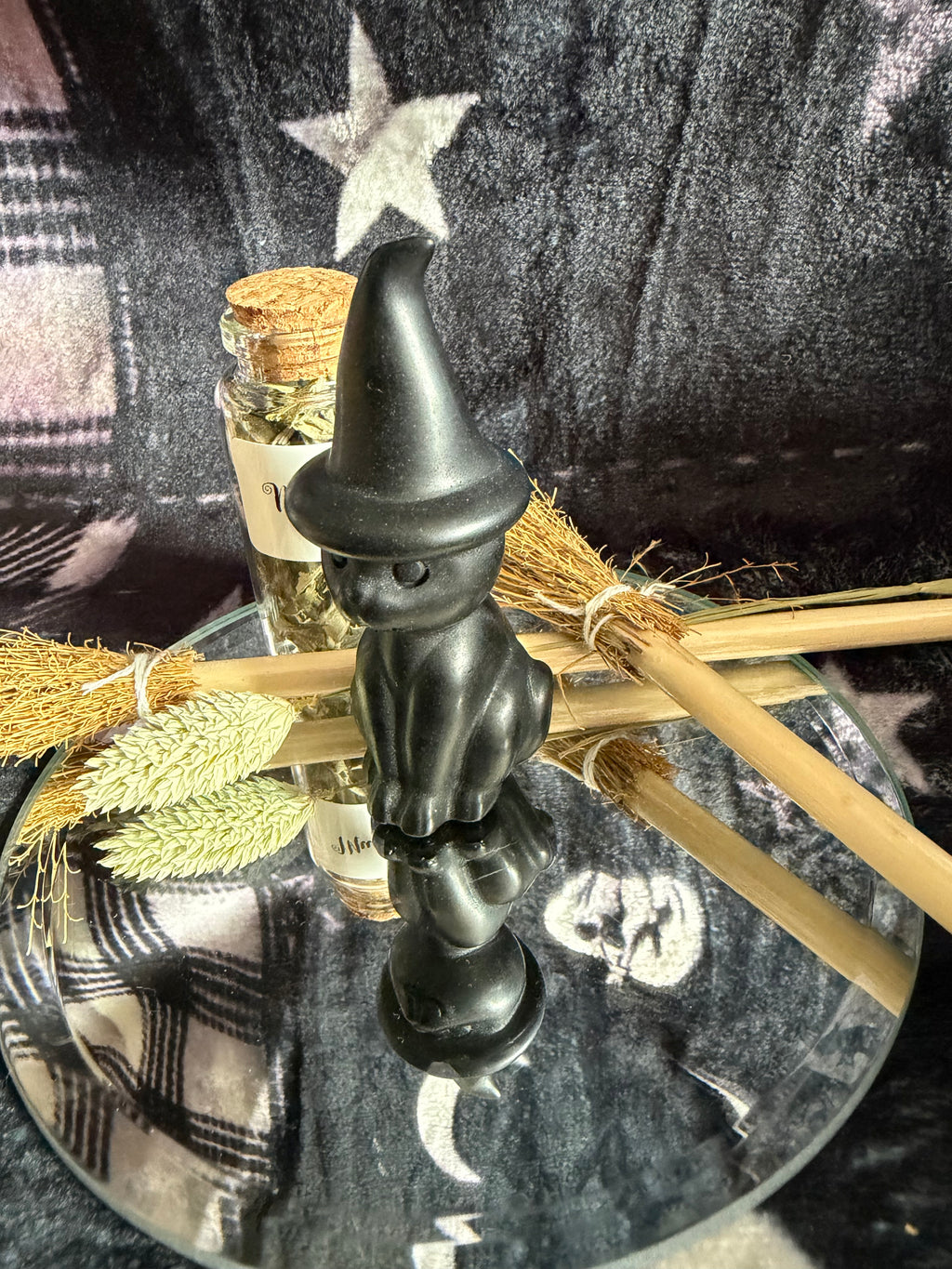 A small black witch figurine with a pointed hat stands on a mirrored tray, surrounded by broomsticks, dried grass, and a small glass vial against a dark plaid fabric background.