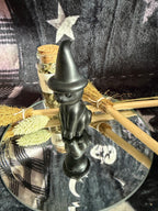 A small black witch figurine with a pointed hat stands on a mirrored tray, surrounded by broomsticks, dried grass, and a small glass vial against a dark plaid fabric background.