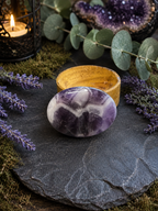 A polished oval purple amethyst crystal with white veining sits on a dark slate tray, styled with a small wooden bowl, eucalyptus leaves, lavender sprigs, moss, and a lit candle in a black lantern.