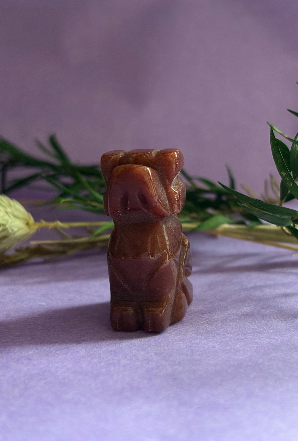 Red Goldstone crystal dog carving front view on lilac background with green leaves — The Crooked Moon