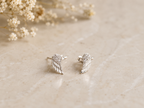 "Angel Wing CZ Stud Earrings Sterling Silver product shot — pair of small silver angel wing stud earrings with pavé cubic zirconia upper wing detail and engraved feather lower wing, on cream marble surface with dried gypsophila"