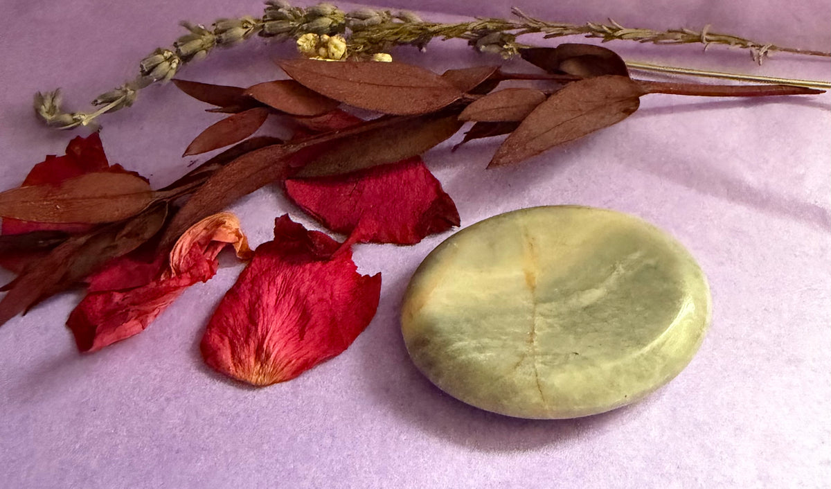 Moonstone palm stone styled on a lavender background alongside dried brown leaves, red rose petals, and a sprig of dried lavender.