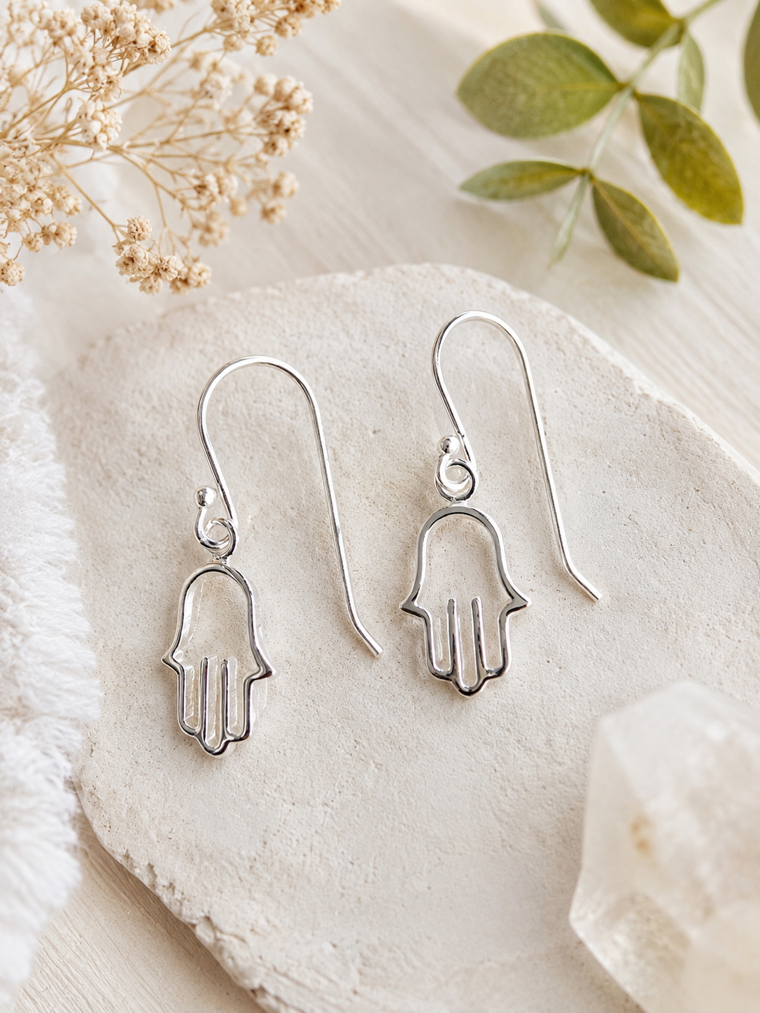 A pair of silver Hamsa hand drop earrings displayed on a textured stone slab with dried flowers and green leaves in the background.
