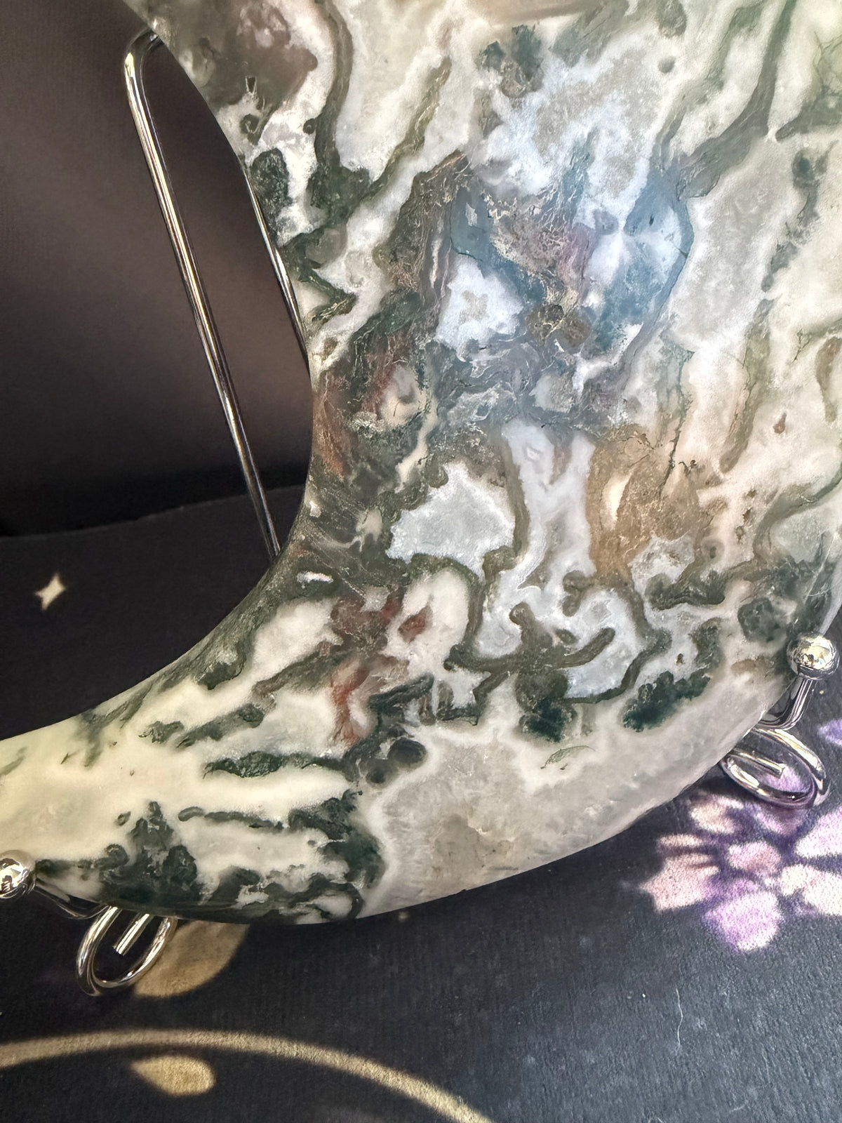 A close-up of a crescent moon-shaped crystal or stone piece with a marbled pattern of white, gray, green, and brown, displayed on a stand.
