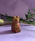 Unakite crystal cat carving on lilac background with green foliage — The Crooked Moon