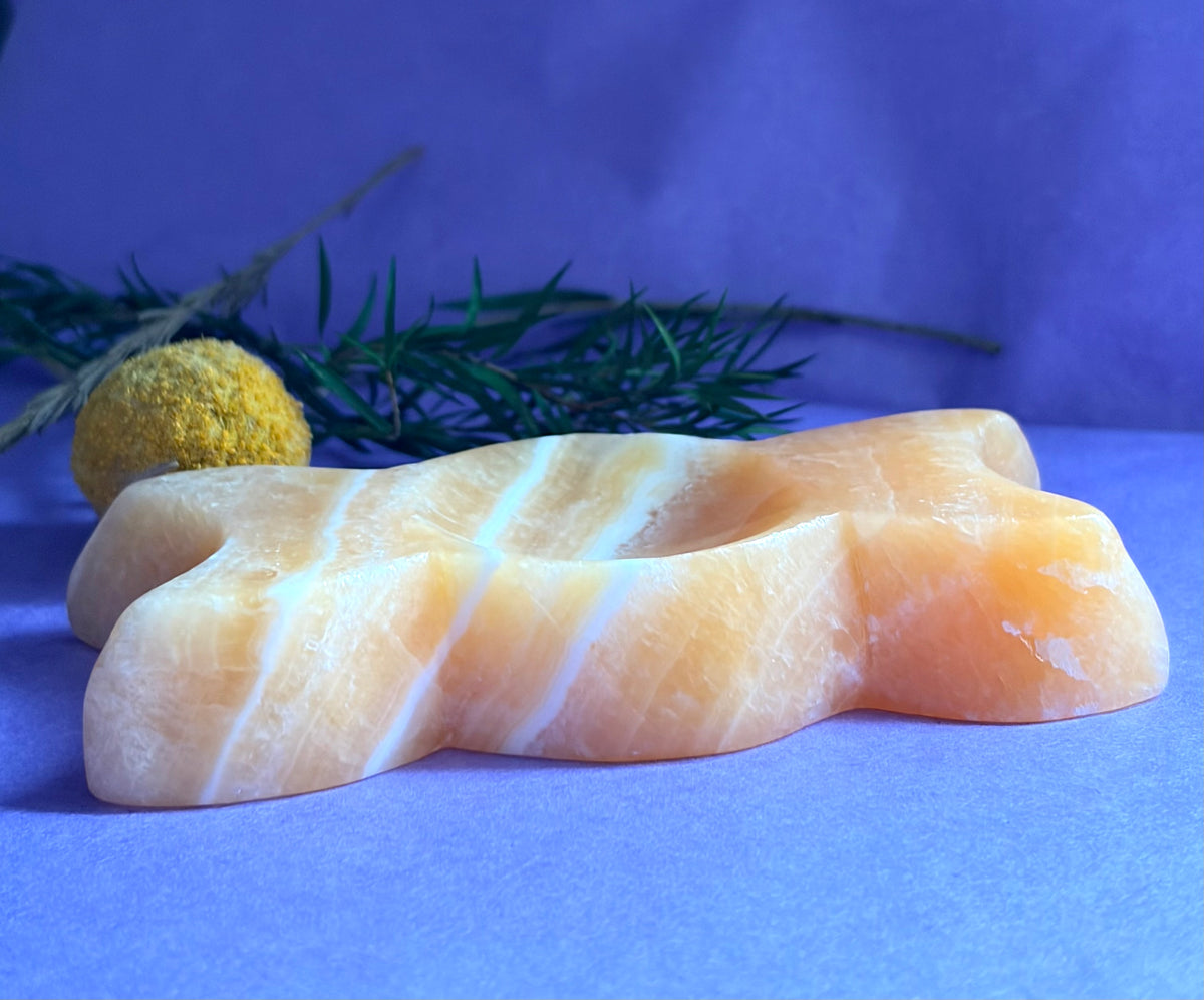 Orange Calcite Triple Moon Goddess Carving displayed on a blue surface with green sprigs and a yellow decorative ball in the background.