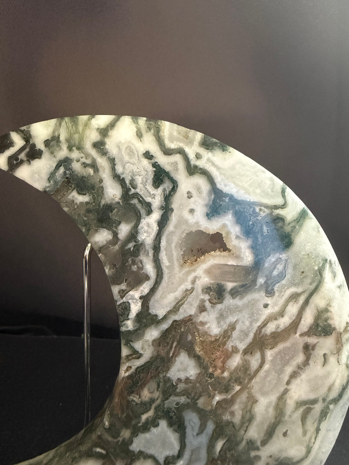 A crescent moon-shaped decorative plaque made of polished agate or marble, featuring swirling patterns of green, gray, white, and blue, displayed on a clear stand against a dark background.