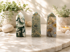 Three polished crystal tower points with natural stone patterns are displayed on a marble slab. The left tower is green and white marbled, the middle is gray with gold leaf, and the right is gray with gold leaf.