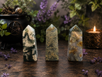 Three polished crystal tower points with natural marbled and metallic patterns, displayed on a rustic wooden surface with a black cauldron, purple flowers, and a burning candle in the background.