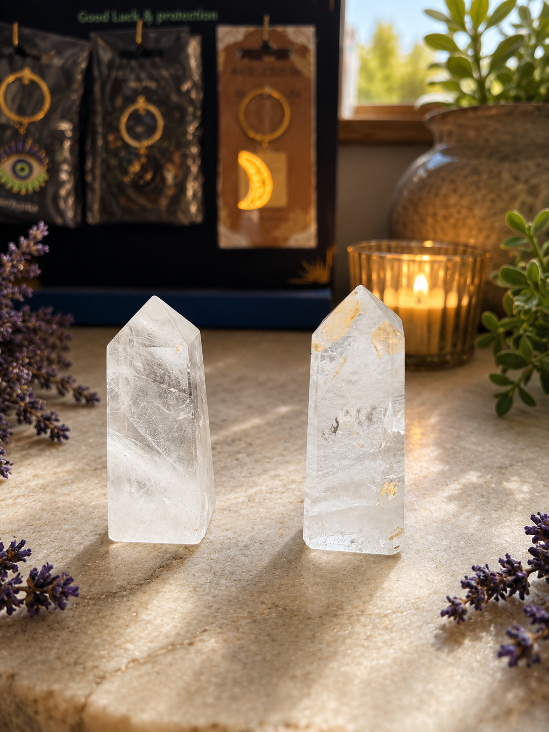 Two clear quartz crystal tower points standing on a stone surface, with a lit candle, plants, and packaged jewelry displayed in the background.