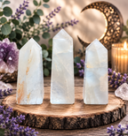 Three tall translucent white crystal towers with natural mineral veining, displayed on a rustic wood slice surrounded by purple flowers, a lit candle, and a decorative crescent moon.