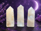 Three pale translucent crystal tower points standing upright on a dark purple surface, with purple flowers and a moon in the background.