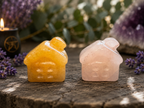 Two small house-shaped crystal carvings, one yellow-orange and one pale pink, displayed on a rustic wooden surface with lavender sprigs, a candle, and a purple geode in the background.