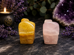 Two small polished stone figurines, one golden yellow and one pale pink, displayed on a dark stone surface with a lit candle, purple flowers, greenery, and an amethyst geode in the background.