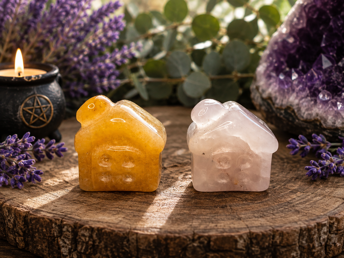 Two small house-shaped crystal stones, one amber-yellow and one pale pink, displayed on a rustic wood slice with lavender, a candle, and an amethyst geode in the background.