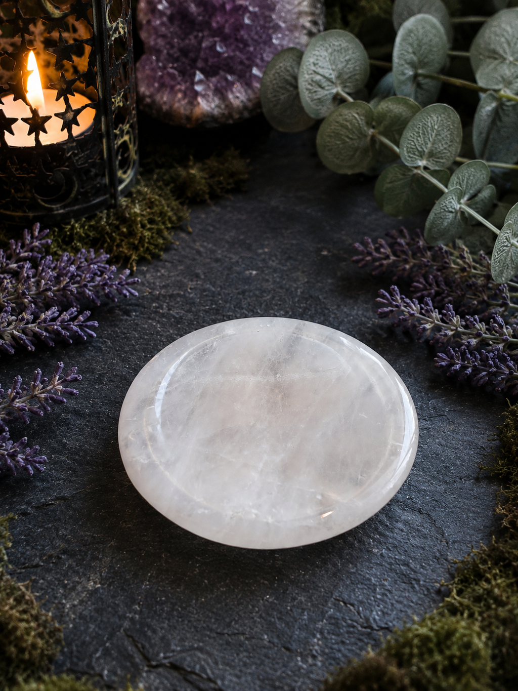A polished white crystal sphere resting on a dark slate surface, surrounded by decorative greenery, purple flowers, moss, and a lit candle lantern.