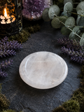 A polished white crystal sphere resting on a dark slate surface, surrounded by decorative greenery, purple flowers, moss, and a lit candle lantern.
