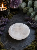 A polished white crystal sphere resting on a dark slate surface, surrounded by decorative greenery, purple flowers, moss, and a lit candle lantern.