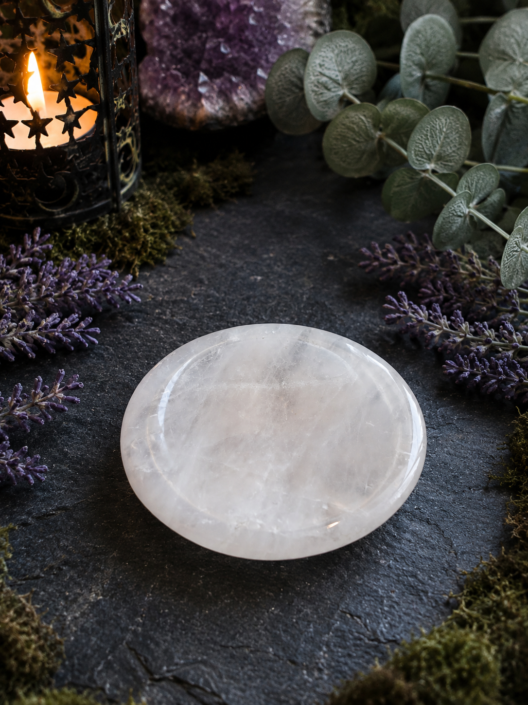 A polished white crystal sphere resting on a dark slate surface, surrounded by decorative greenery, purple flowers, moss, and a lit candle lantern.