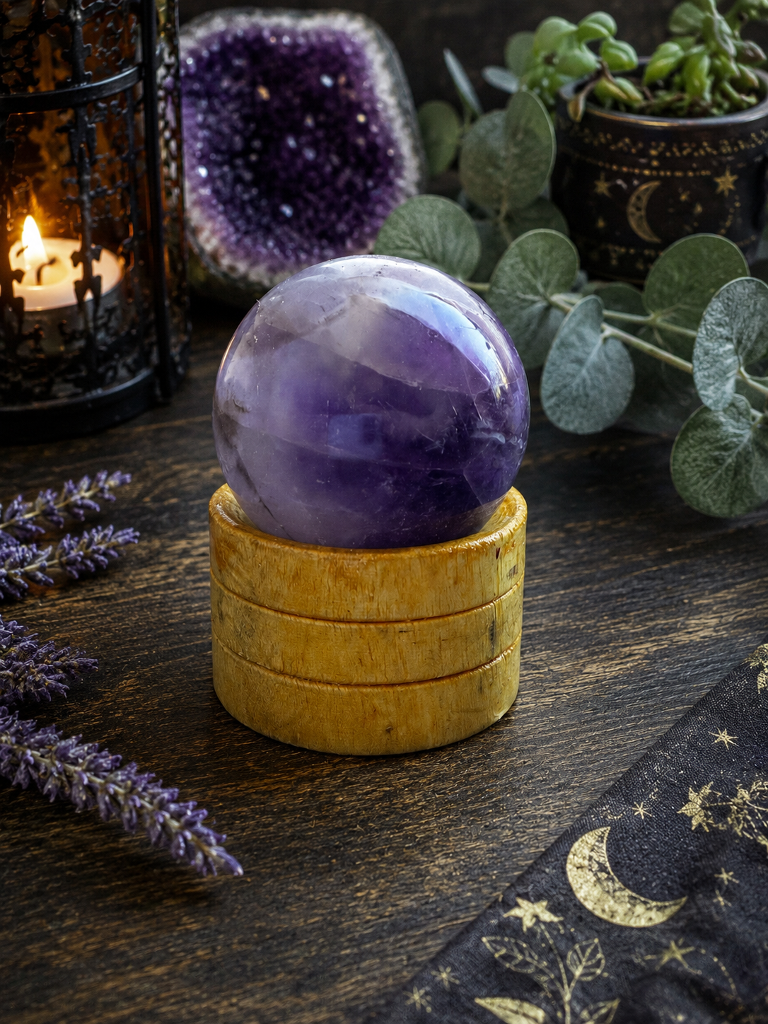 A polished purple amethyst crystal sphere resting on a stack of three light wood coasters, styled on a dark wooden table with a candle, amethyst geode, eucalyptus leaves, and a starry fabric napkin.