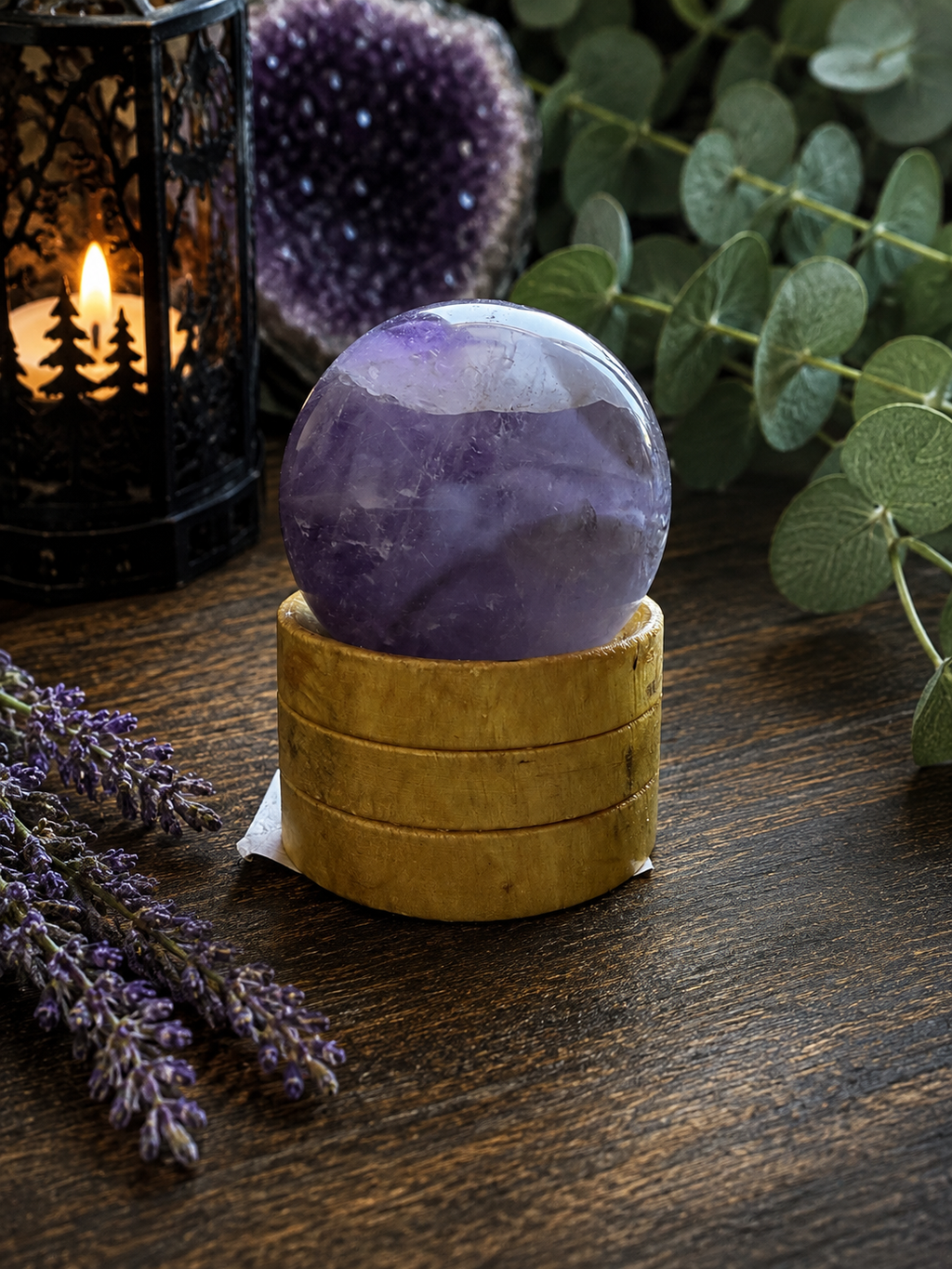 A polished purple amethyst crystal sphere resting on a small round wooden stand, set on a dark wooden tabletop with decorative greenery, lavender, and a lit black lantern in the background.