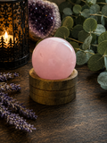 A polished pink rose quartz crystal sphere resting on a small wooden stand, styled on a dark wooden surface with a lit candle lantern, purple lavender sprigs, a geode, and green eucalyptus leaves.