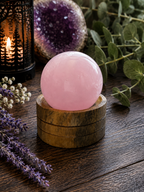 A polished pink rose quartz crystal ball resting on a small round wooden base, styled on a dark wooden table with dried lavender, green leaves, a lit lantern, and a purple crystal geode in the background.