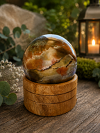 A polished agate crystal sphere with natural earthy patterns sits on a round wooden stand, placed on a rustic wooden surface with eucalyptus leaves, a blurred stone, and a glowing lantern in the background.