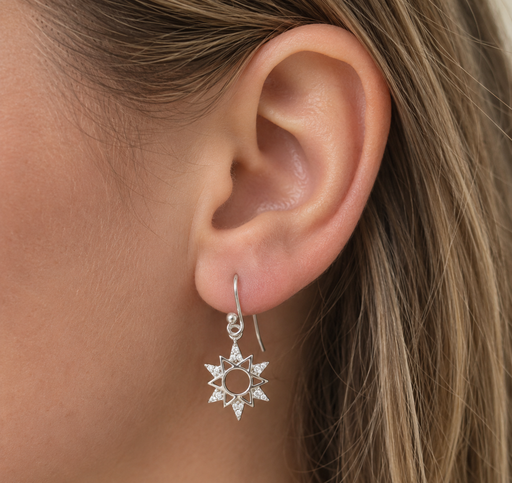 A close-up of a person's ear wearing a silver starburst dangle earring with small clear stones, against a background of light brown hair.