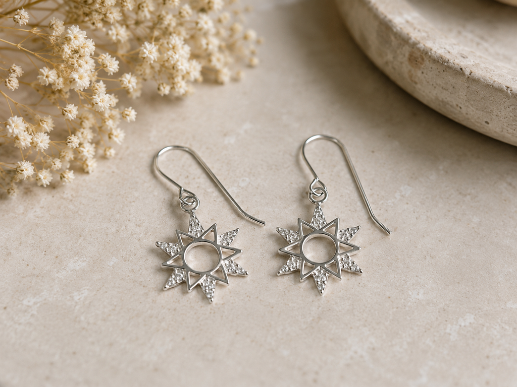 A pair of silver dangle earrings with starburst sun motifs, displayed on a textured beige surface next to dried white flowers.