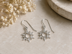 A pair of silver dangle earrings with starburst sun motifs, displayed on a textured beige surface next to dried white flowers.