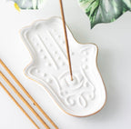 Hamsa Hand Incense Stick Holder lifestyle shot with incense stick burning on white surface with tropical leaves
