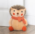 Hazel The Hedgehog plush doorstop propped against white door on wooden floor lifestyle shot