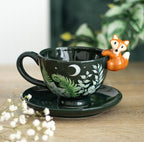 Dark green ceramic fox tea cup and saucer with botanical fern design, fox figurine on rim, lifestyle shot with gypsophila flowers