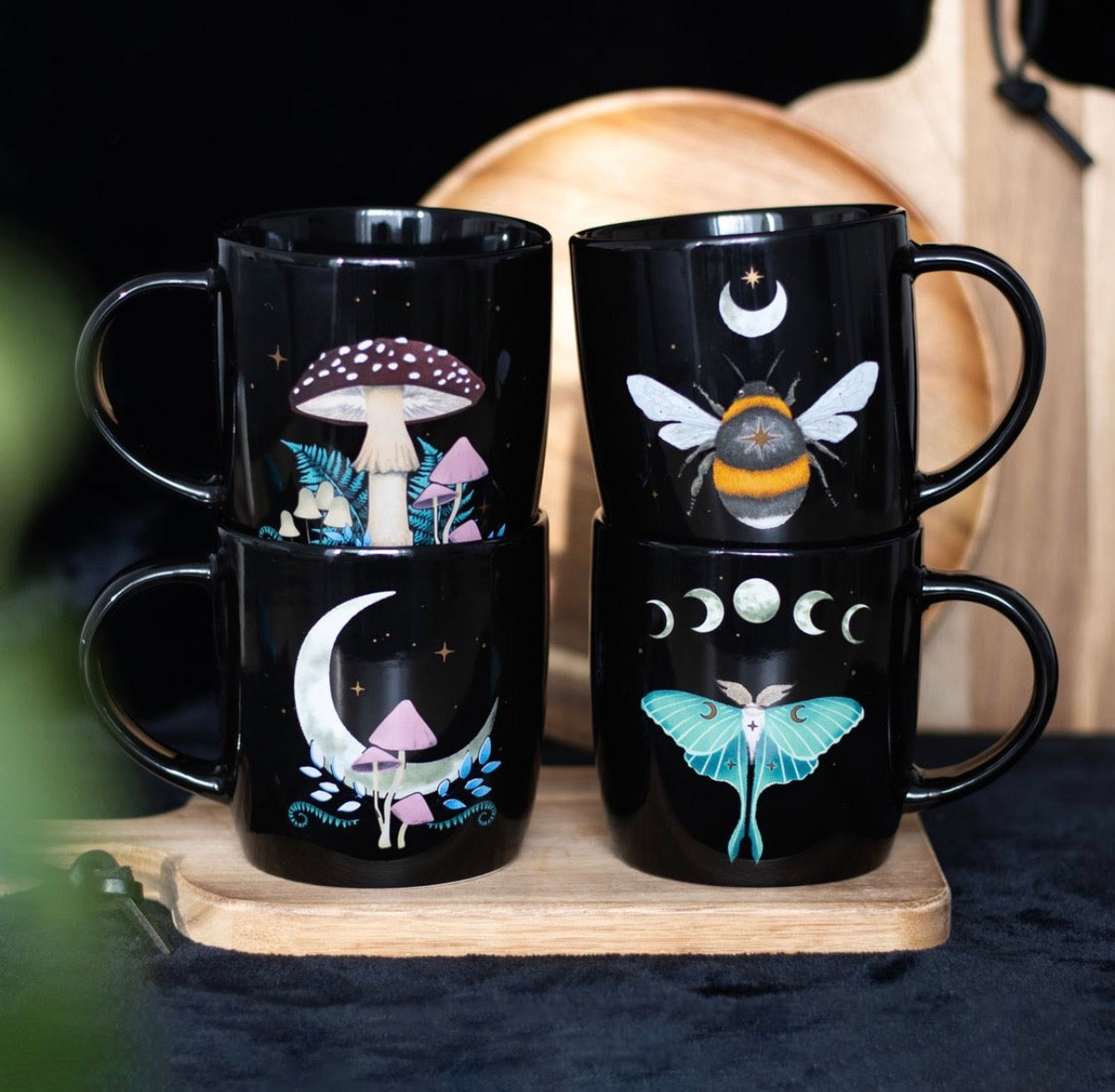 Four black Dark Forest mugs stacked in pairs on a wooden board, showing mushroom, moon mushroom, celestial bee and luna moth designs, dark moody lifestyle shot