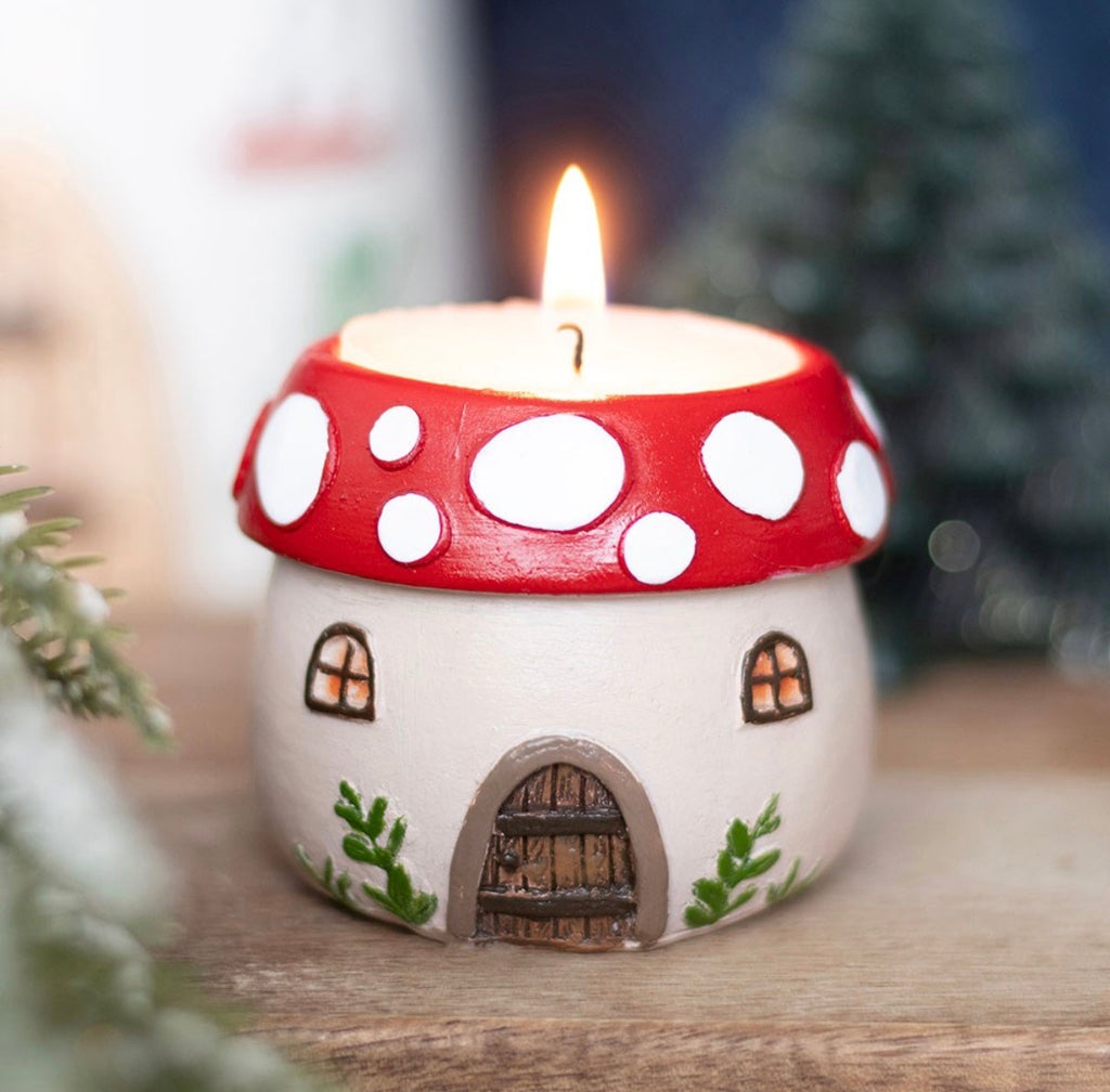 Mushroom house tealight holder lit with candle flame, red spotted cap and painted door and windows, moody lifestyle shot with pine branches