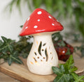Small ceramic mushroom tealight holder lit with warm candlelight glowing through cut-out botanical stem design, red spotted cap, surrounded by ivy leaves on wooden board