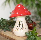 Small ceramic mushroom tealight holder lit with warm candlelight glowing through cut-out botanical stem design, red spotted cap, surrounded by ivy leaves on wooden board