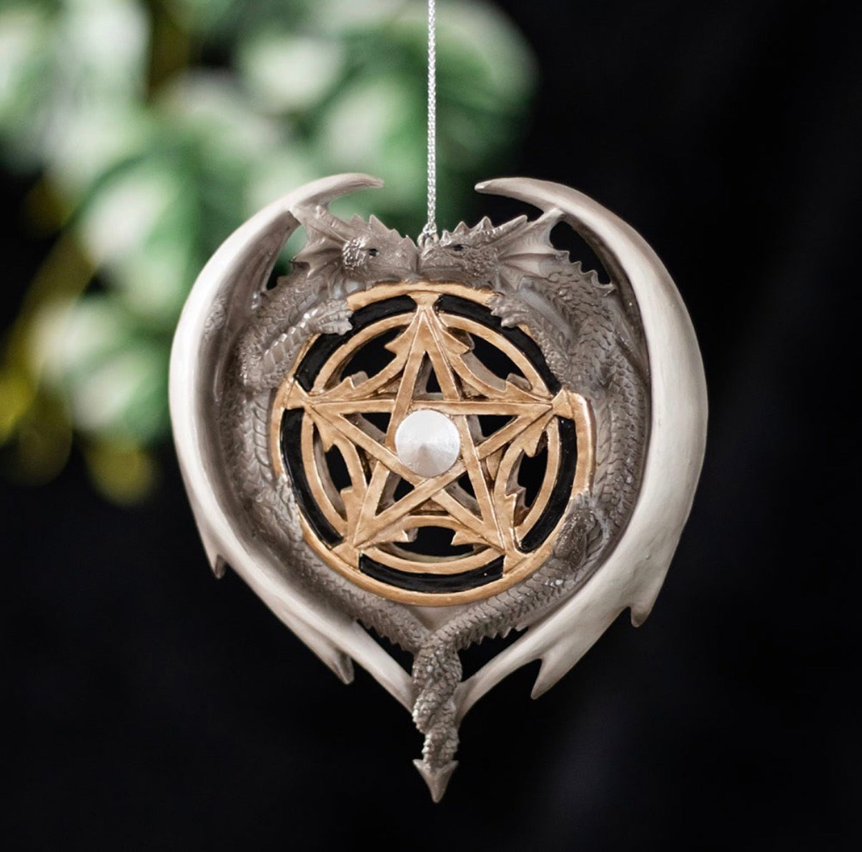 Anne Stokes Dragon Magic hanging ornament showing two entwined silver dragons forming a heart around a gold pentagram with pearlescent gem, dark background with foliage