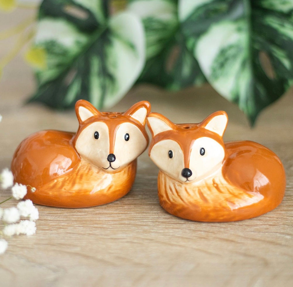 Two ceramic fox salt and pepper shakers in russet and cream facing each other on a wooden surface with foliage in background