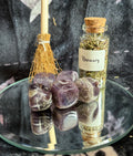 A glass jar labeled 'Rosemary' filled with dried herbs sits on a round mirrored tray beside polished purple stones and a small straw broom against a dark textured backdrop.